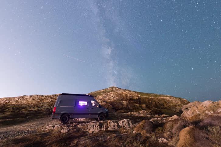 A gray camper van sits on a rocky ridge under a vibrant milky way as light glows from a side window against the dark mountain landscape and star filled sky