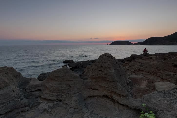 Rocks and mountains along the coast as a fisherman sits near the water and the sun sets on the horizon