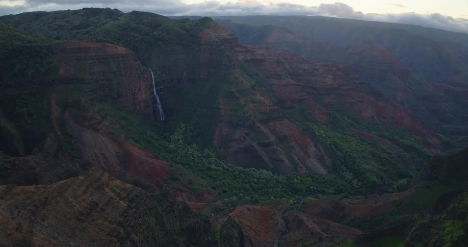 A stunning aerial view slowly pushes in on the majestic Waimea Canyon in Hawaii, revealing a beautiful waterfall cascading down the lush green and red rock walls at dusk