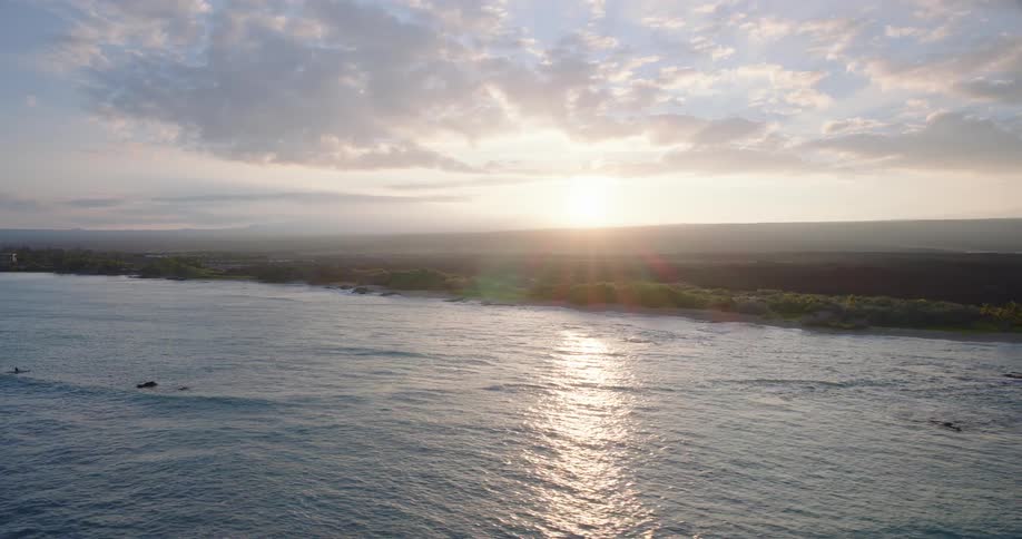 An aerial view of the sun rising over a tropical coastline, with its light reflecting on the calm ocean waves