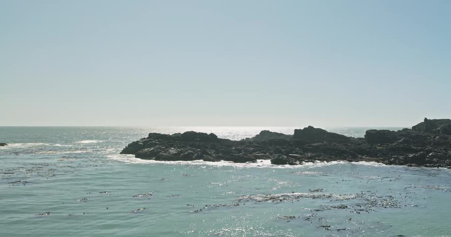 Slow zoom towards a rugged, rocky coastline as tranquil ocean waves lap the shore on a sunny day