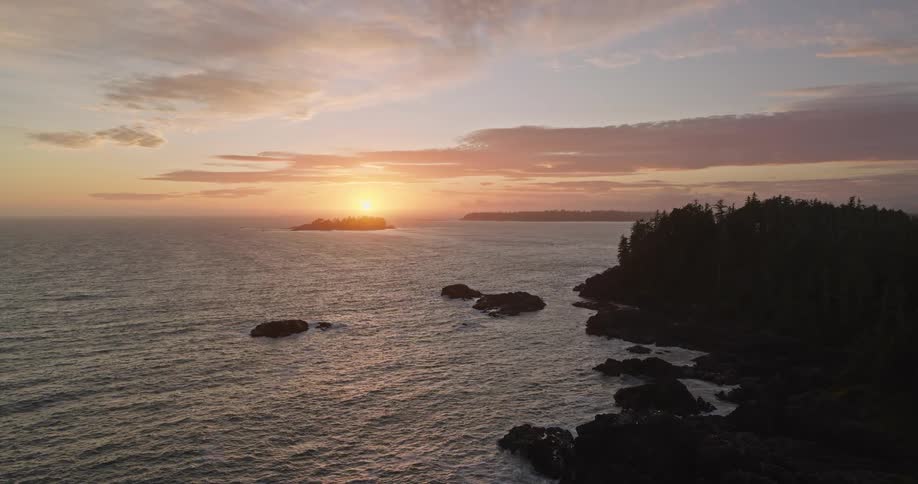 An aerial view captures the sun setting over the tranquil ocean along a rugged, tree-lined coastline