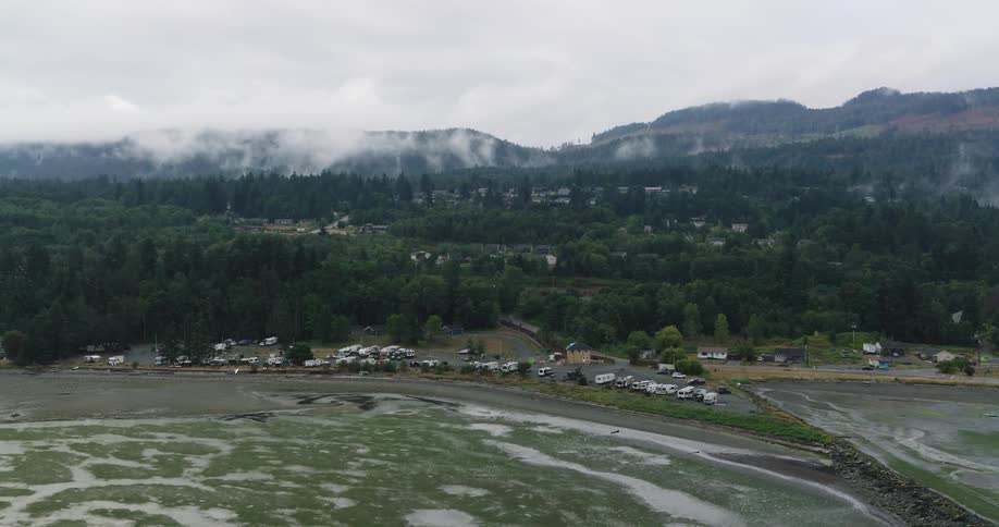 A static aerial view of an RV park on a misty coastline, with dense forests and foggy mountains in the background during low tide