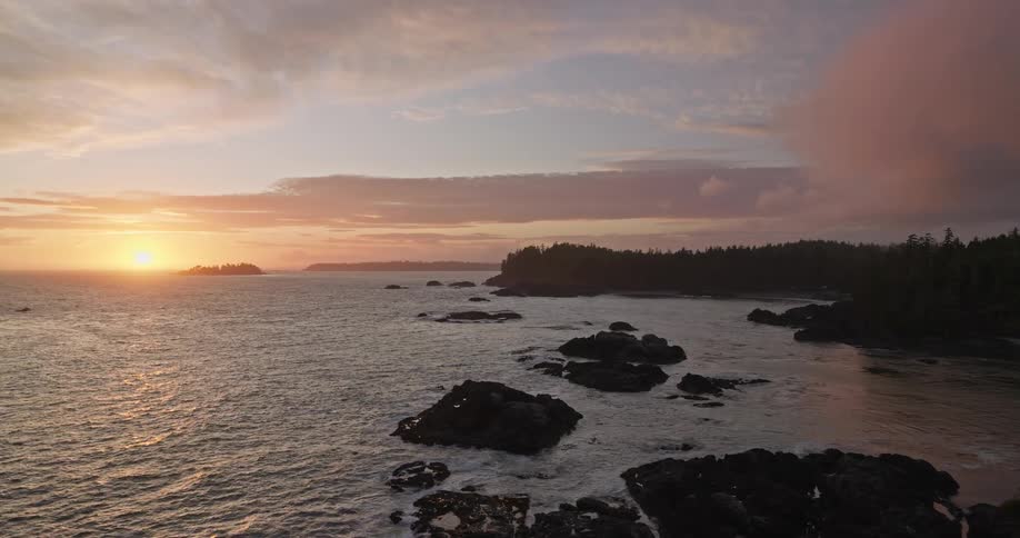 A stunning aerial view shows the sun setting over a rugged, tree-lined coastline as gentle waves wash over the dark rocks
