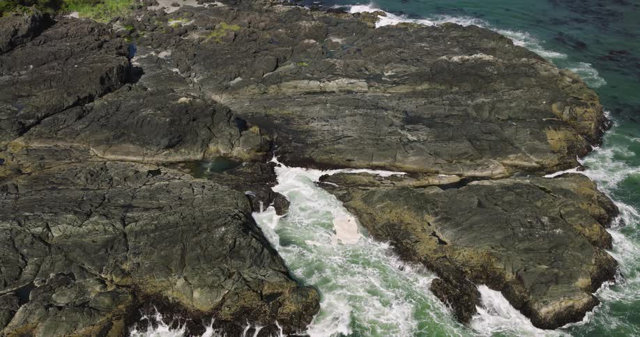From high above, the camera slowly descends toward a rocky shoreline where green ocean water furiously crashes and foams