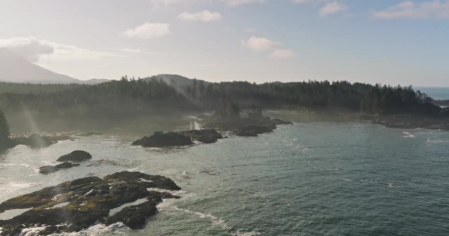 An ethereal aerial view of a rugged, misty coastline where sun rays pierce through the haze over the ocean and a dense evergreen forest
