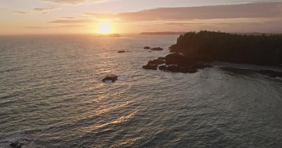 An awe-inspiring aerial view reveals a rugged, forested coastline as the golden sun sets over the tranquil ocean