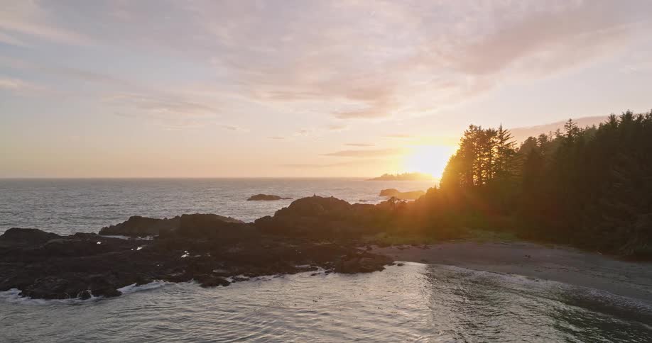 A serene aerial shot captures the sun setting over a rocky Pacific Northwest coastline, illuminating the tranquil ocean and lush forest