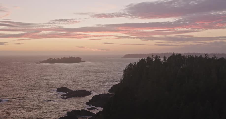 An aerial view pulls back to reveal a rugged, tree-lined coastline with rocky shores as the sun sets over the vast ocean