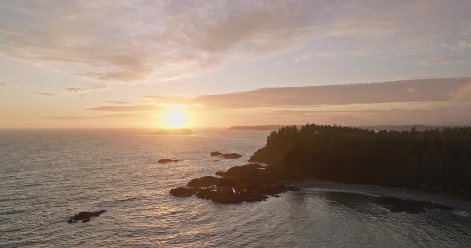 Aerial view of a rugged, tree-lined coast with rocky shores as the sun sets beautifully over the Pacific Ocean