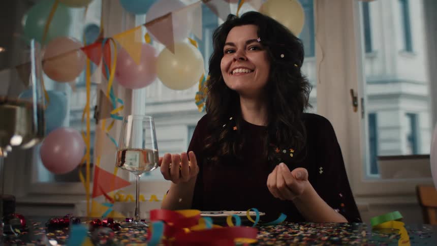 Joyful woman blows a handful of colorful confetti towards the camera in slow motion during a party