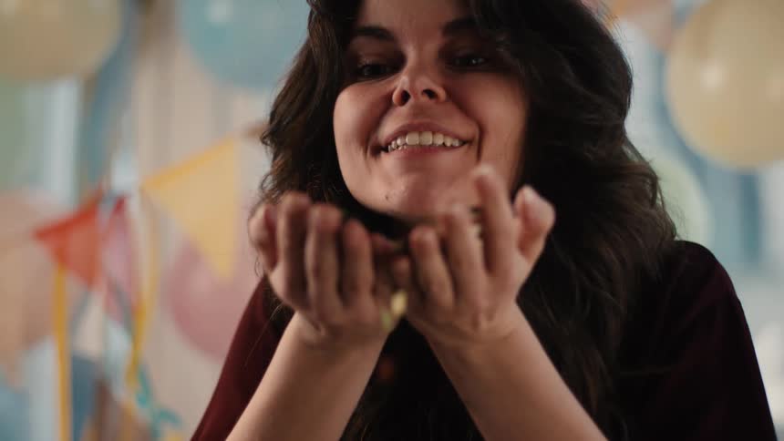 A joyous woman blows a handful of colorful confetti towards the camera in slow motion during a festive celebration