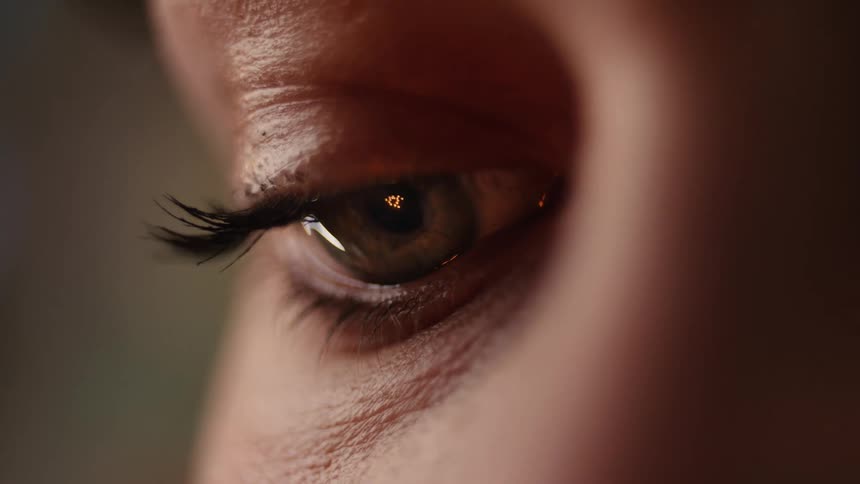 An extreme close-up macro shot of a woman's eye with a warm, flickering light reflecting in her pupil