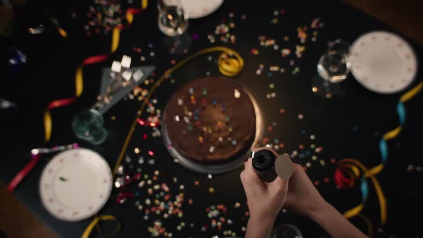A top-down view shows hands popping a confetti cannon over a chocolate birthday cake on a decorated party table in super slow motion