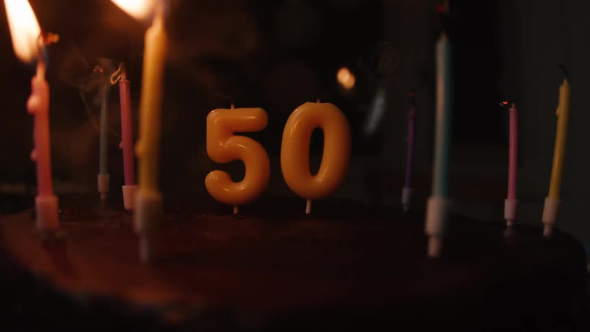 Candles on a 50th birthday cake are blown out one by one, leaving trails of smoke in the darkness