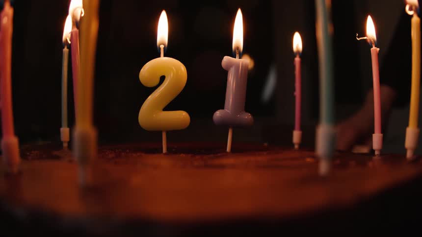 A person lights the candles shaped like the number 21 on a chocolate birthday cake in a dark room