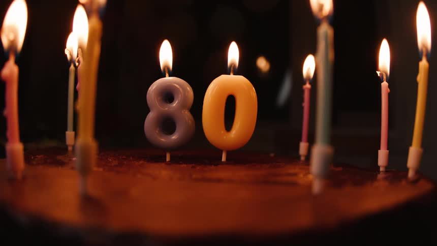 A birthday cake with lit candles forming the number 80 glows in a dark room