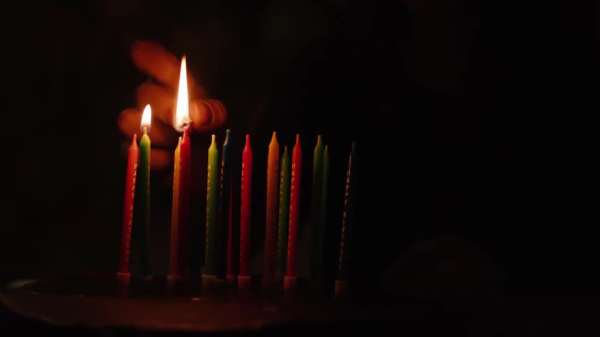 In a dark room, a hand uses one lit candle to light a row of colorful birthday candles in slow motion