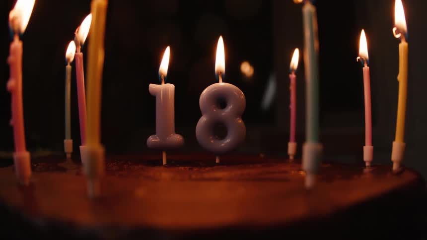 A hand carefully lights the number 18 candles on a chocolate birthday cake, surrounded by smaller colorful candles, in a dimly lit room