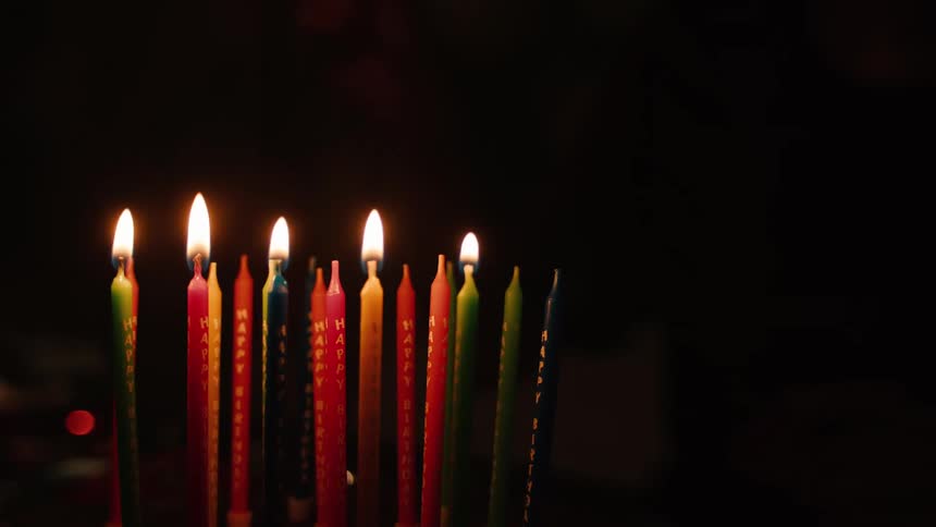 A hand slowly lights colorful 'Happy Birthday' candles with a lighter against a dark background