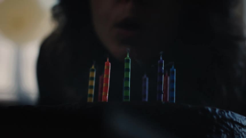A person blows out colorful birthday candles on a dark chocolate cake in dramatic slow motion