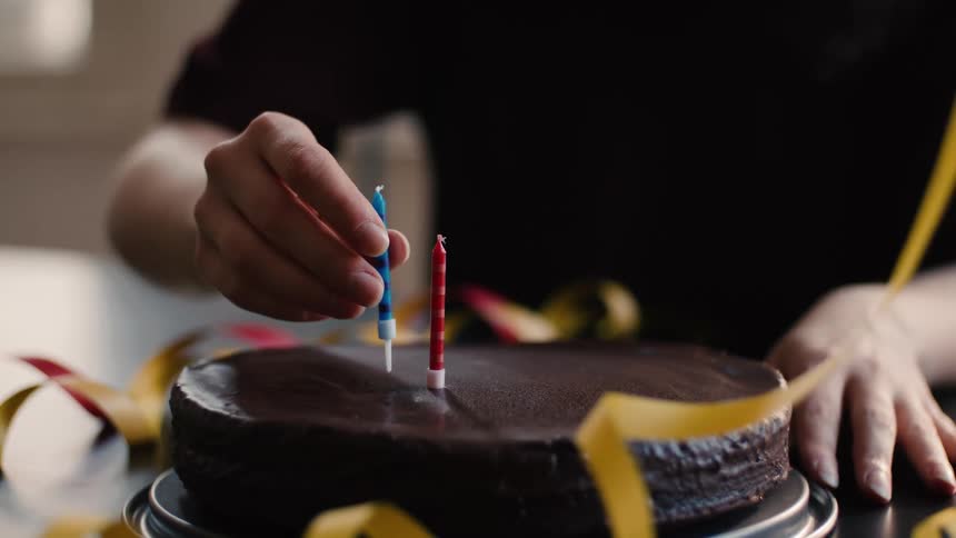 A person carefully places two colorful birthday candles, one by one, into a rich chocolate cake in slow motion