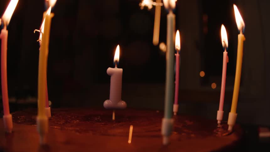 A number one candle burns brightly on a birthday cake surrounded by colorful candles and festive sparklers in a dark room
