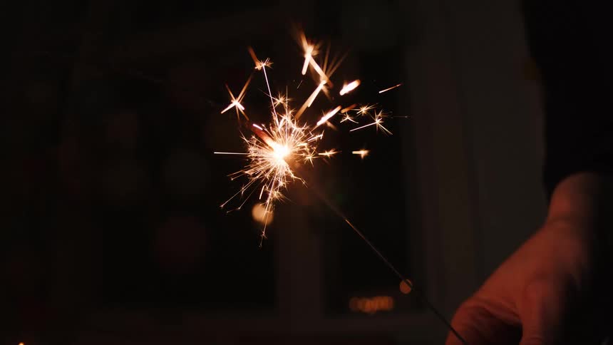 A hand holds a brightly burning sparkler in slow motion against a dark, blurry background, creating a mesmerizing display of glowing sparks