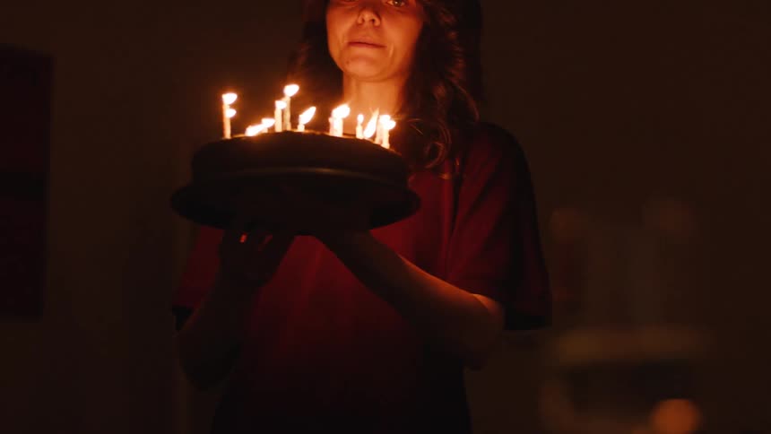 A woman slowly walks out of a dark hallway into a room, holding a birthday cake illuminated by its lit candles