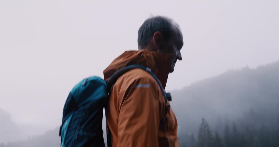 A determined older man with a backpack walks along a wet road through a foggy, atmospheric landscape in slow motion