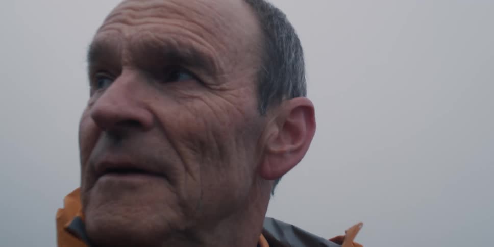 A thoughtful elderly man in an orange jacket looks off into the distance on a foggy day