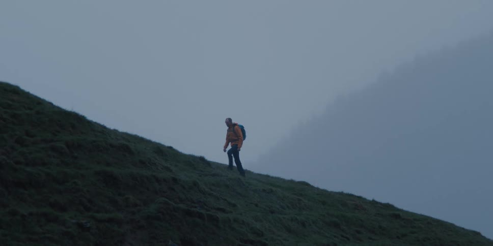A lone hiker in a bright jacket slowly ascends a steep, grassy mountain through dense fog in slow motion