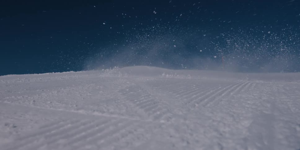 A lone skier in a green jacket carves down a groomed mountain slope in slow motion, kicking up a spray of snow against the deep blue sky