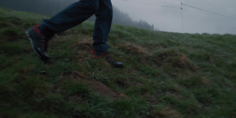 A lone hiker in an orange jacket and backpack walks in slow motion down a grassy slope on a foggy, overcast day