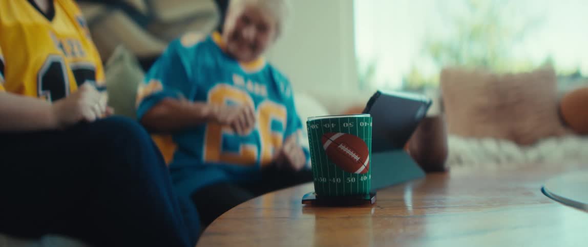 A football-themed drink koozie sits on a coffee table as the focus shifts to an older and younger woman in jerseys excitedly watching a game on a tablet