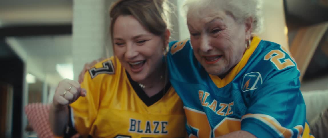 A young woman and an older woman in football jerseys cheer and laugh with excitement while watching a game together at home in slow motion