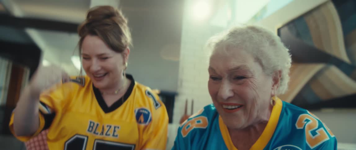 An elderly woman and a young woman in football jerseys laugh together while watching a game at home
