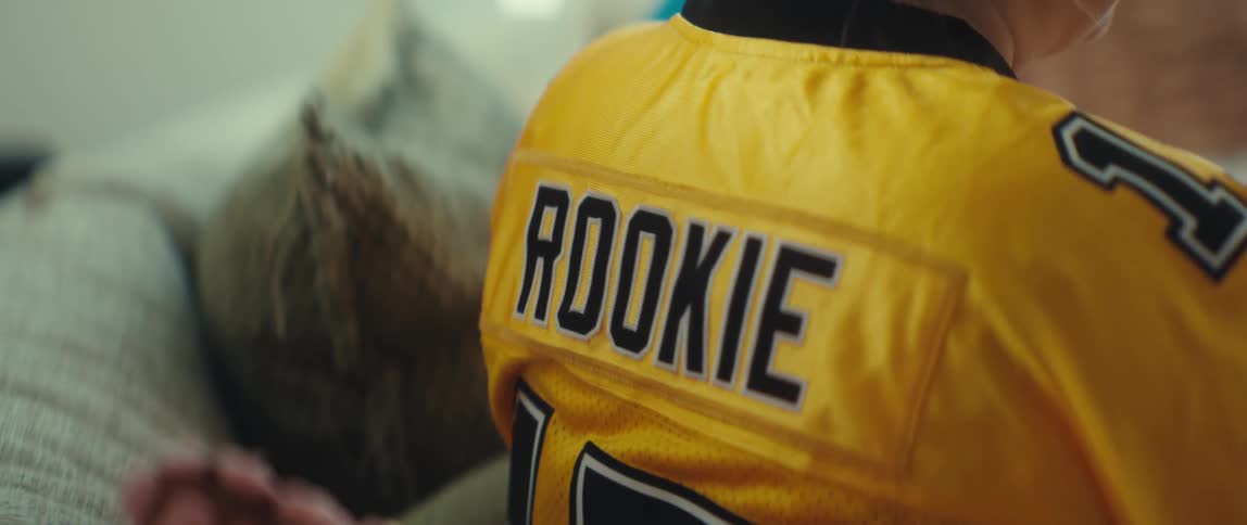 An over-the-shoulder close-up of a fan wearing a yellow 'Rookie' football jersey while watching a game with family