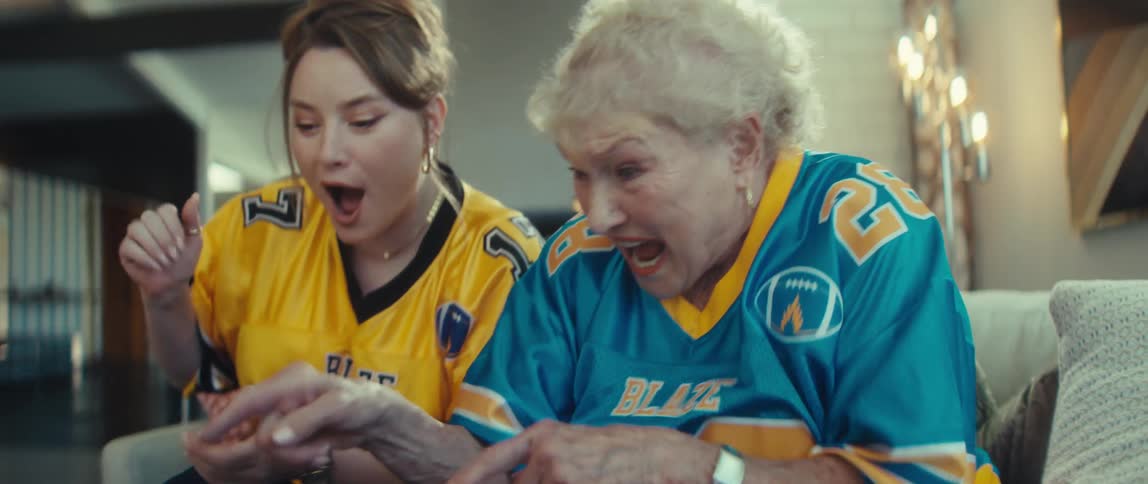 A young woman and her grandmother celebrate excitedly while watching a football game together on a tablet from their couch