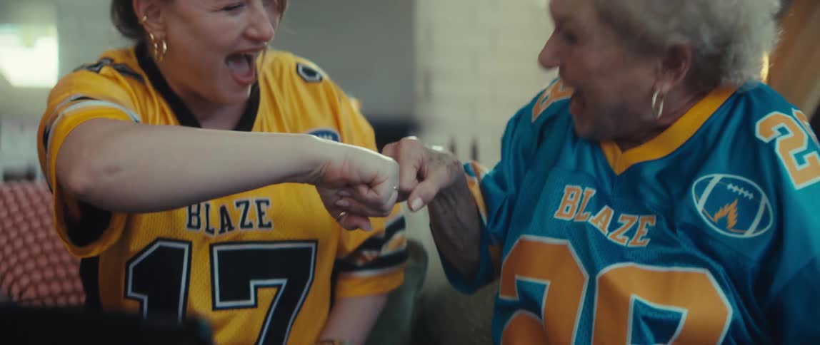 An excited young woman and her grandmother celebrate with a slow-motion fist bump while watching a football game on a tablet
