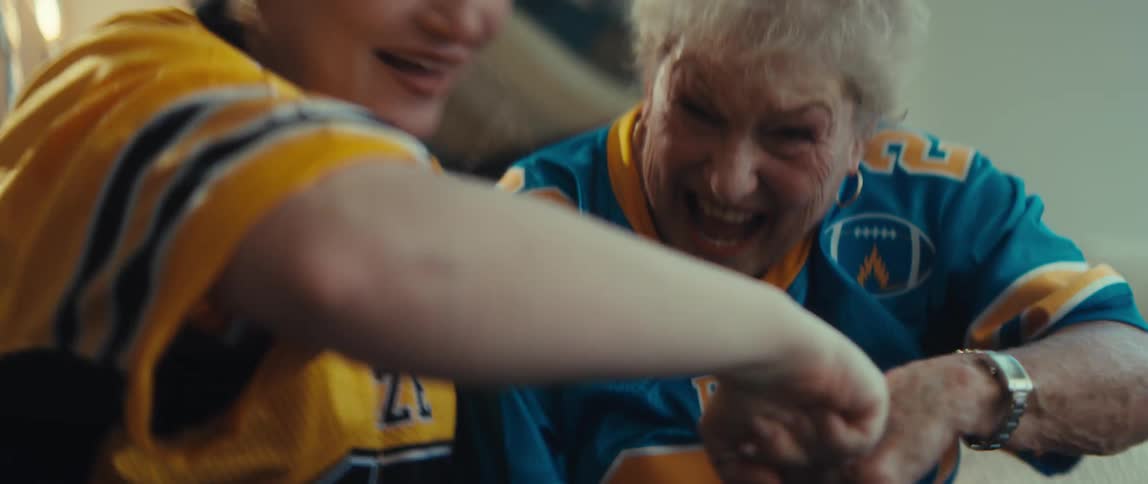 An exuberant grandmother and granddaughter, wearing football jerseys, cheer and fist-bump while watching a game together at home in slow motion