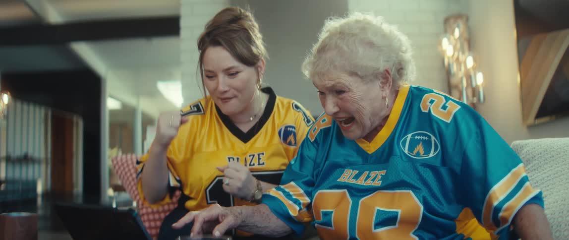 Two excited women in football jerseys cheer and dance in slow motion while winning a fantasy football game on a tablet