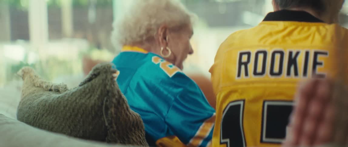 Close up from behind of two football fans in bright jerseys sitting on a couch, with 'ROOKIE' and the number 17 on one shirt