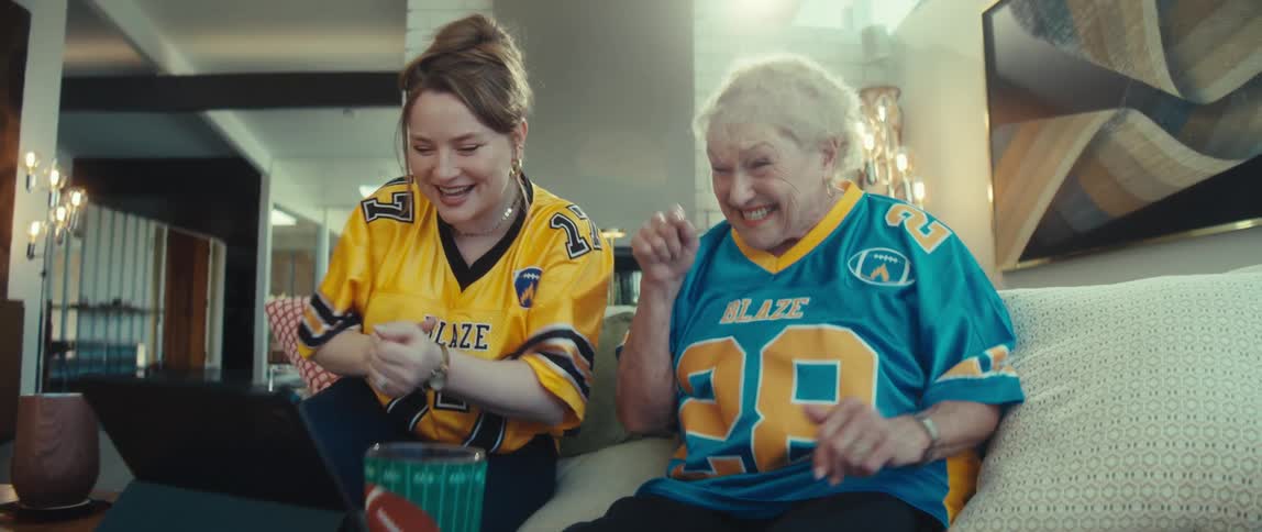 An excited granddaughter and her grandmother wearing matching football jerseys cheer, dance, and fist bump while watching a game on a tablet at home