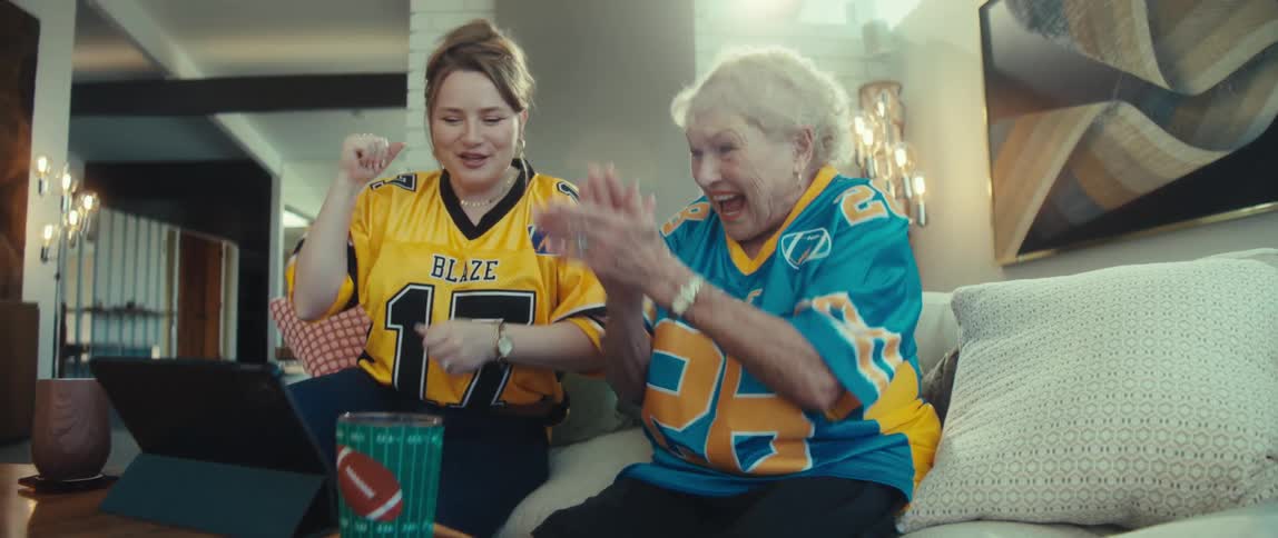 An excited young woman and her grandmother in opposing football jerseys cheer and dance while watching a game on a tablet