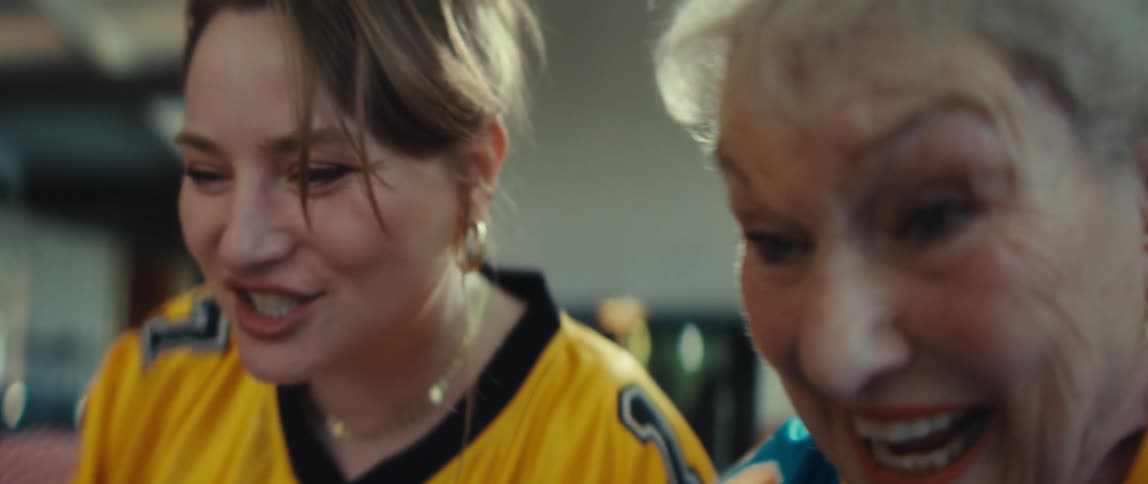 An excited young woman in a football jersey watches a game with intense anticipation before exploding with joy and celebration alongside her elderly friend
