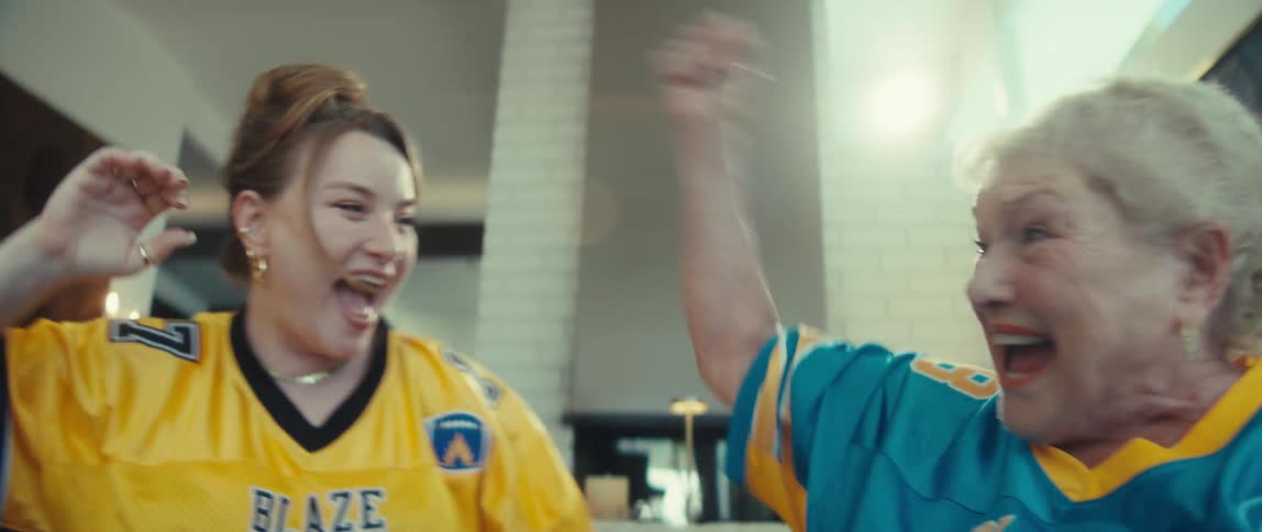 A young woman and her grandmother wearing football jerseys cheer, high-five, and celebrate in slow motion while watching a game