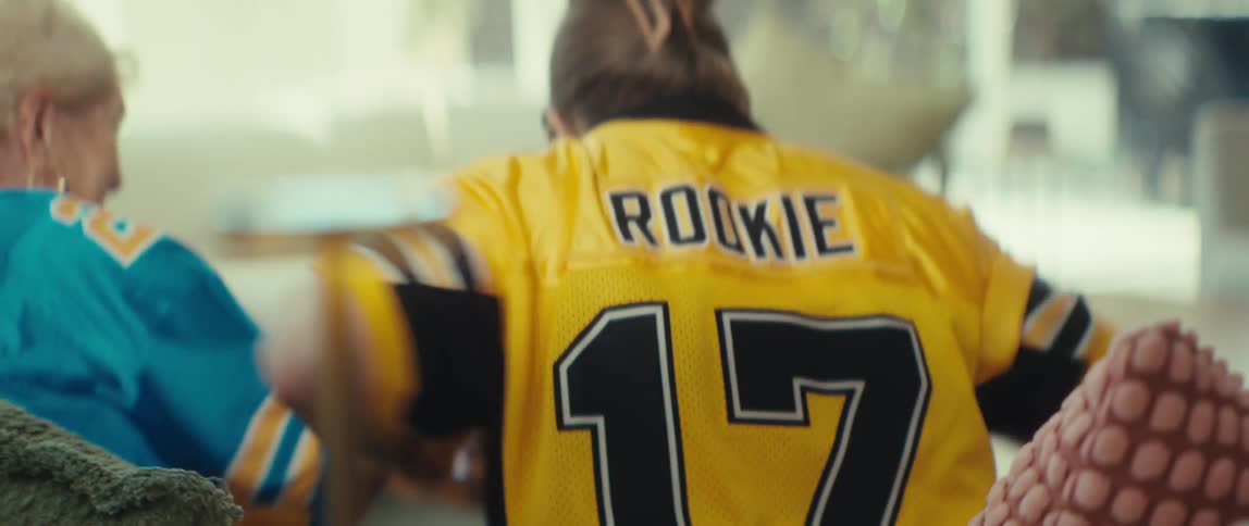 An elderly woman wearing a 'Legend' jersey and a younger woman in a 'Rookie' jersey cheer for their team while watching a game at home