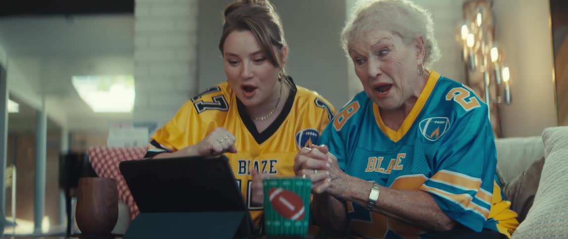 A granddaughter and her grandmother, wearing football jerseys, share a moment of pure excitement as they watch the game and celebrate together on a tablet