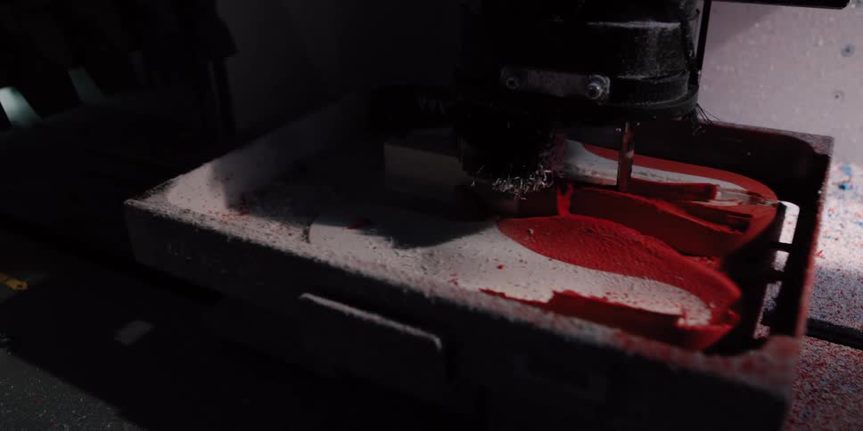 An industrial CNC milling machine meticulously carves a custom shape from a layered red and white block in a dimly lit workshop